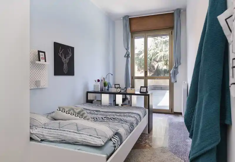 Chambre lumineuse et chaleureuse avec un lit simple bien fait, coussins décoratifs et un bureau près d'une grande fenêtre qui inonde la pièce de lumière naturelle.