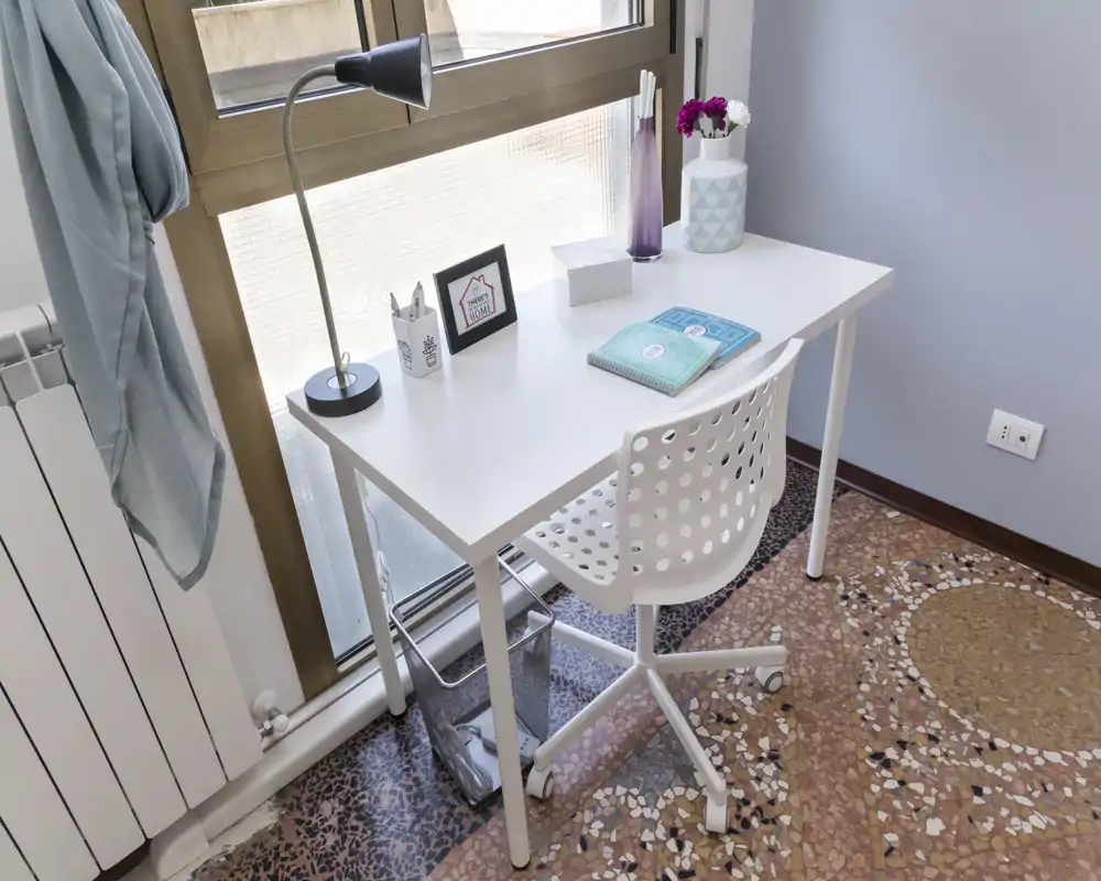 Angolo studio luminoso e ordinato vicino a una grande finestra con scrivania bianca minimal, sedia traforata e pochi accessori decorativi — perfetto per lavoro da remoto o studio.