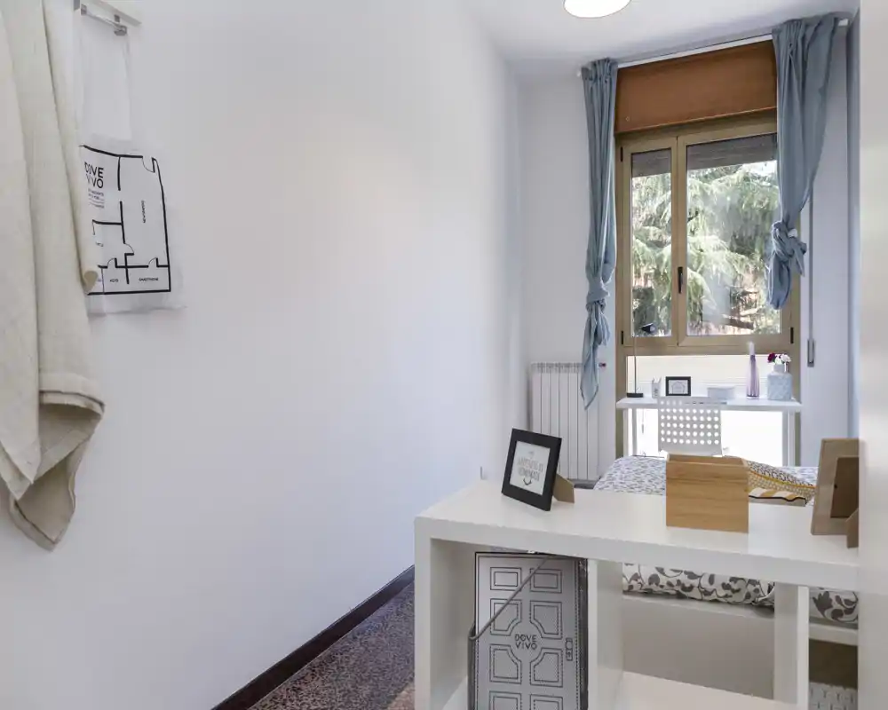 Camera singola luminosa e minimale con scrittoio vicino alla finestra. La stanza presenta tessili morbidi, un piccolo scaffale ordinato e luce naturale dalla finestra ampia.