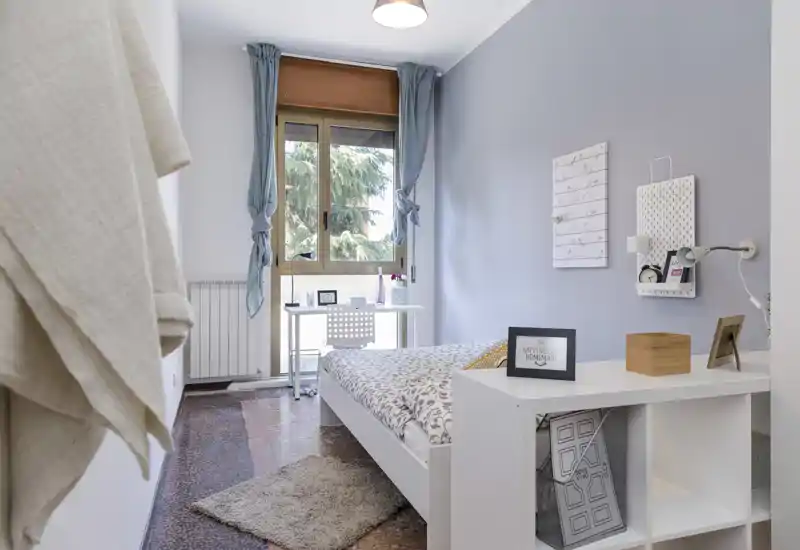 Bright, modern single bedroom with a white bed, desk by the window and soft decorative accents, ideal for a cozy rental.