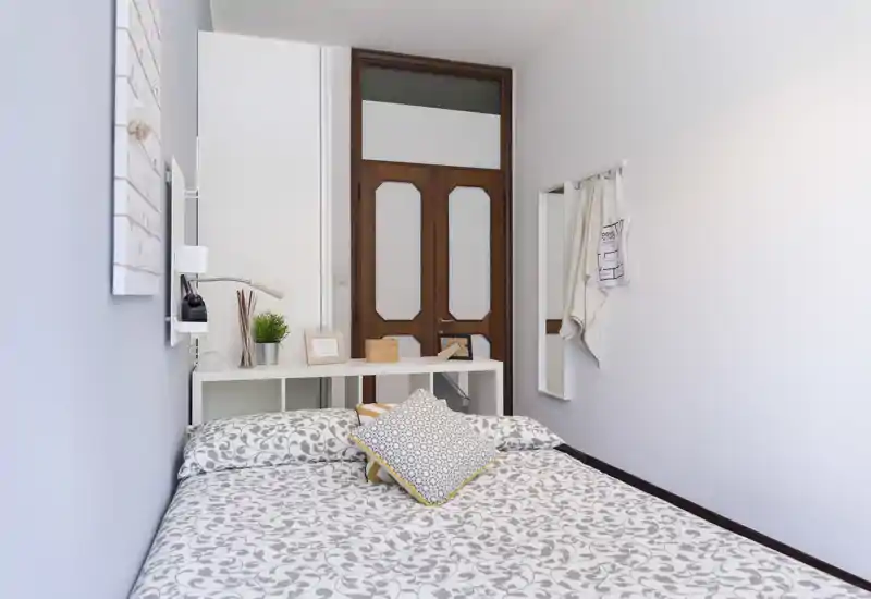 Bright, tidy bedroom with a patterned double bed, decorative cushions and a slim console behind the headboard; neutral walls and wooden door add warmth—ideal for a short-stay listing.