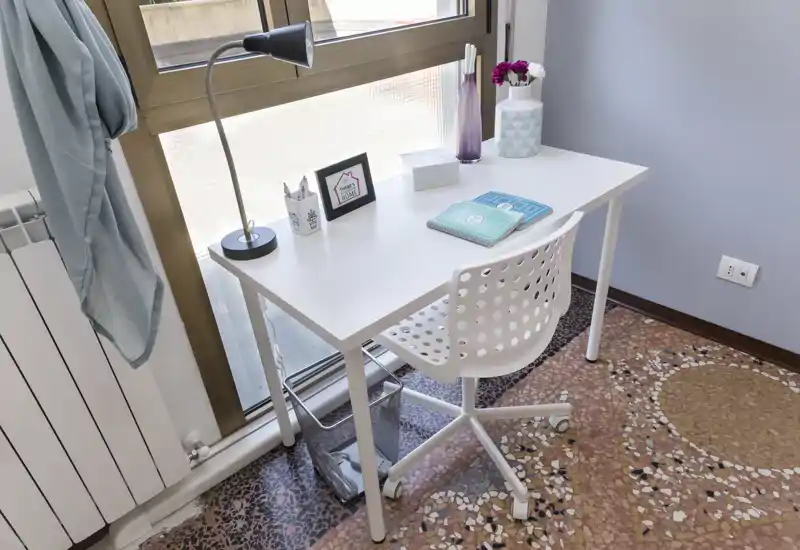 Angolo studio luminoso e ordinato vicino a una grande finestra con scrivania bianca minimal, sedia traforata e pochi accessori decorativi — perfetto per lavoro da remoto o studio.