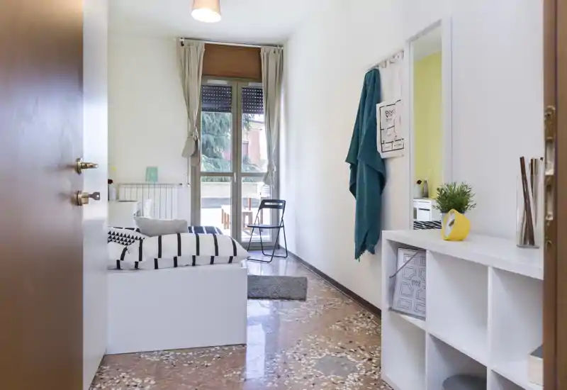 Camera singola luminosa e ordinata con letto a righe, luce naturale dalla porta finestra e una pratica libreria all'ingresso — ideale per uno studente o inquilino singolo.
