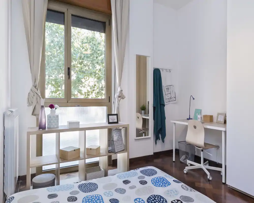 Chambre lumineuse et soignée avec couette à motifs, grande fenêtre et bureau—idéale pour un étudiant ou un professionnel recherchant un espace calme.