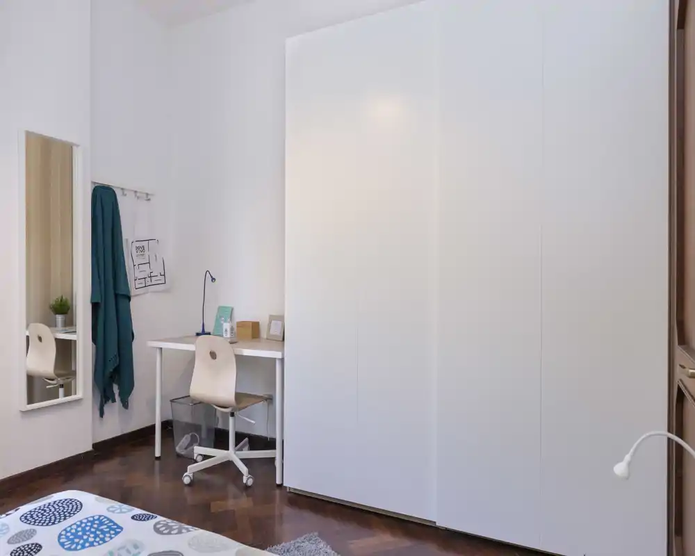 Coin chambre lumineux et minimaliste avec une grande armoire blanche, un petit bureau et une partie du lit avec une couette à motifs. Style propre et moderne, avec un espace de travail pratique, idéal pour se reposer ou étudier.