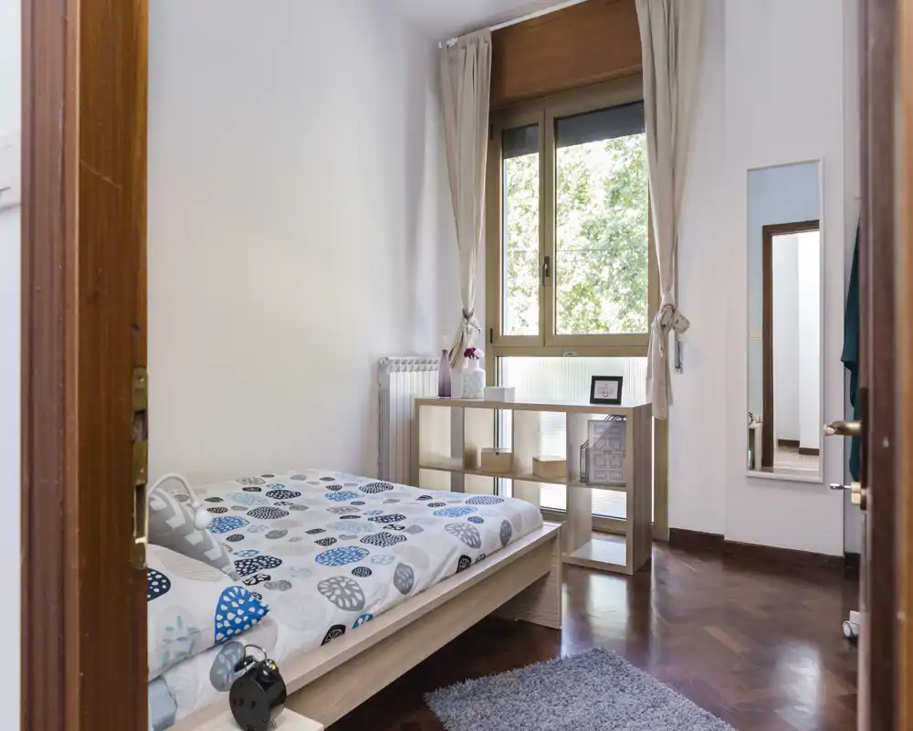 Chambre lumineuse et épurée avec lit double, parquet et grande fenêtre apportant une belle lumière naturelle. Tons neutres, petit meuble de rangement et textiles doux pour une atmosphère apaisante.