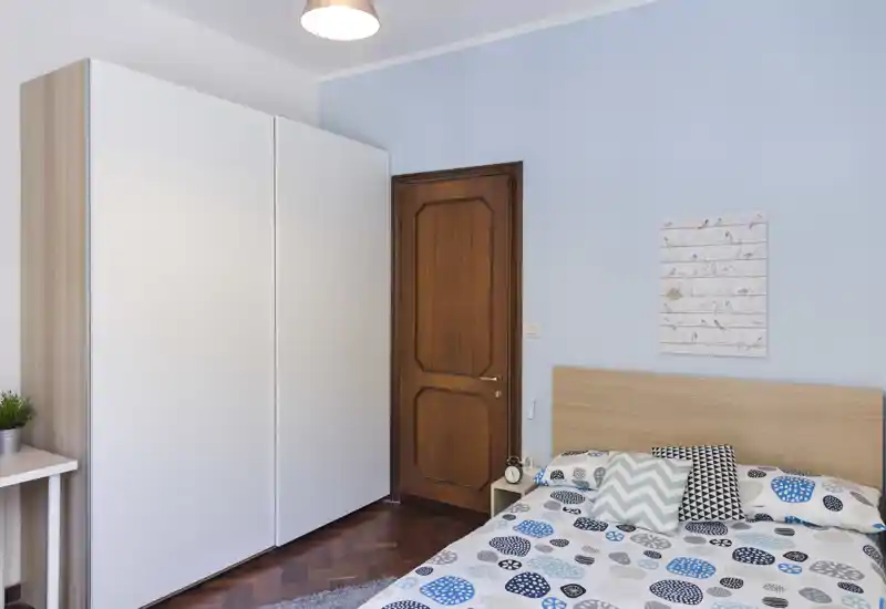 Chambre lumineuse et bien rangée avec tête de lit en bois, linge de lit à motifs et une grande armoire blanche. L'espace paraît frais et accueillant grâce à une douce lumière naturelle et des coussins décoratifs modernes.