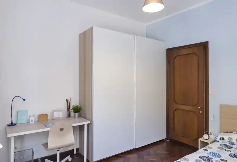Coin chambre propre et lumineux avec armoire, petit bureau et lit partiellement visible avec couverture à motifs — idéal pour une personne ou un étudiant.