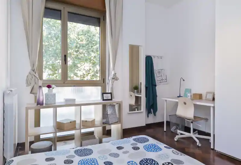 Chambre lumineuse et soignée avec couette à motifs, grande fenêtre et bureau—idéale pour un étudiant ou un professionnel recherchant un espace calme.