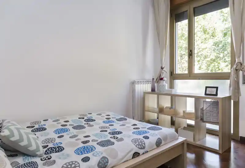 Chambre lumineuse et minimaliste avec un lit double à motif, une étagère simple près d'une grande fenêtre et un parquet chaleureux — idéale pour un séjour cosy et lumineux.