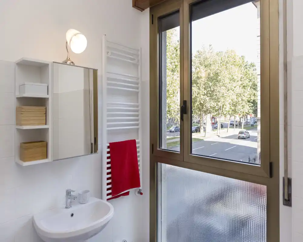 Bagno luminoso e ben curato con lavabo a parete, pensile con specchio, termoarredo e una grande finestra che offre luce naturale e vista sulla strada.