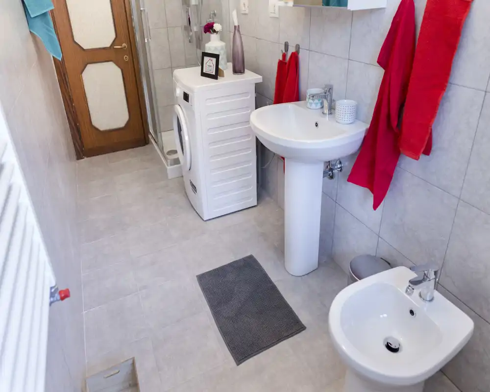 Bagno moderno e pulito con lavabo a colonna, bidet, cabina doccia e lavatrice; piastrelle neutre e illuminazione chiara rendono l'ambiente ordinato.