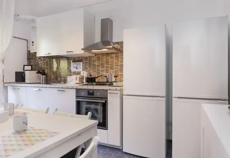 Bright modern kitchen with white cabinetry, a built-in oven, gas hob and two tall white refrigerators; a small dining table with placemats completes the scene.