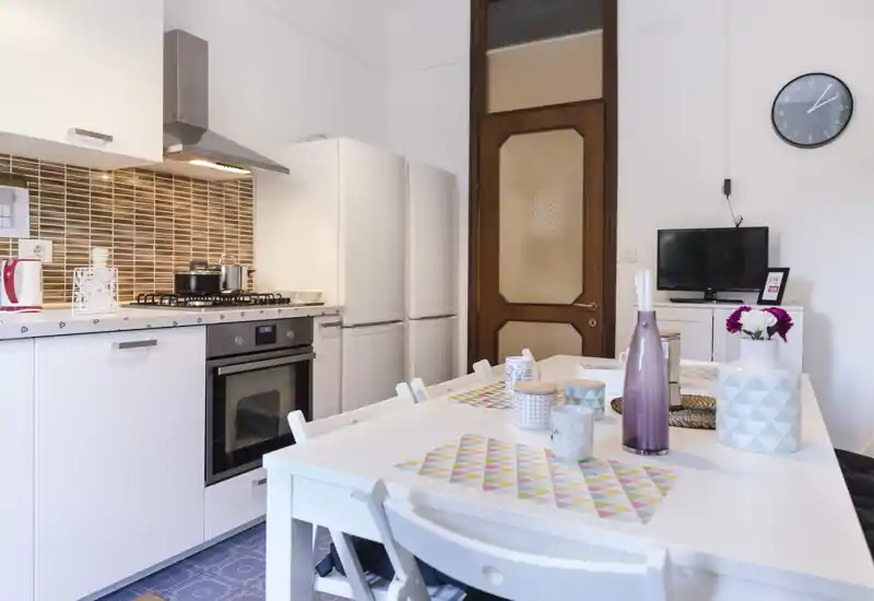 Bright, modern kitchen with white cabinetry, built-in oven, refrigerator and a dining table set with decorative vases and placemats — ideal for home rental listings.