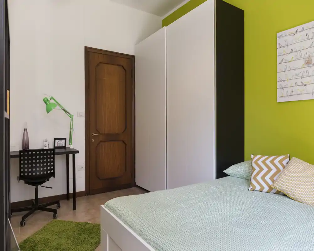 Comfortable, brightly lit bedroom with a green accent wall, a bed dressed in soft textiles and decorative cushions, a large white wardrobe and a compact study desk — ideal for a single occupant.