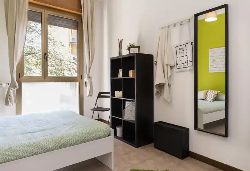Bright, tidy bedroom with a double bed dressed in green linens, a shelving unit and a full-length mirror reflecting the room for added depth.