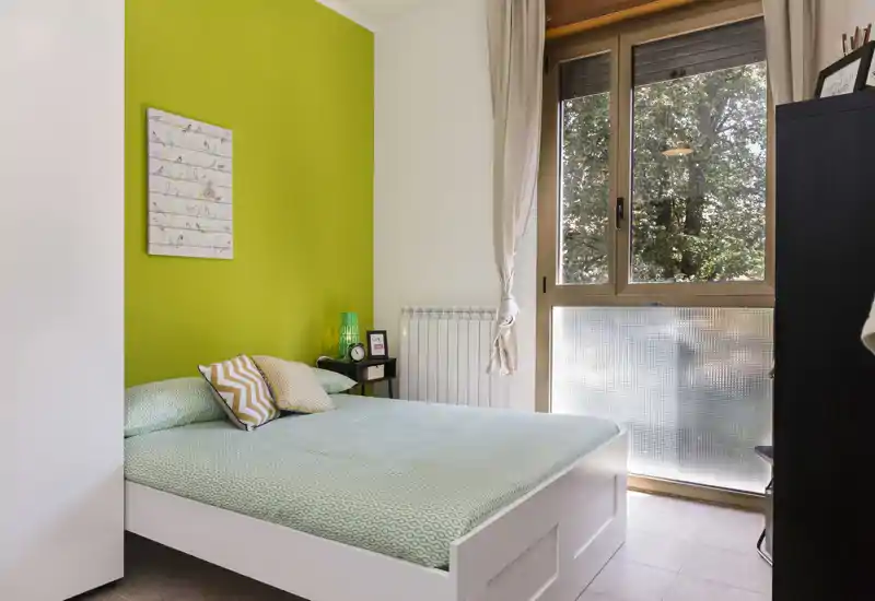 Sunlit bedroom with a green accent wall, full-size bed with patterned bedding, bedside table and large window letting in natural light — calm and fresh atmosphere.