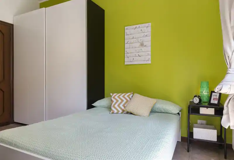 Bright, modern bedroom with a double bed dressed in soft green linens, decorative pillows and a bedside table with lamp and alarm clock. A white wardrobe and a vivid green accent wall complete the cozy, well-styled space.