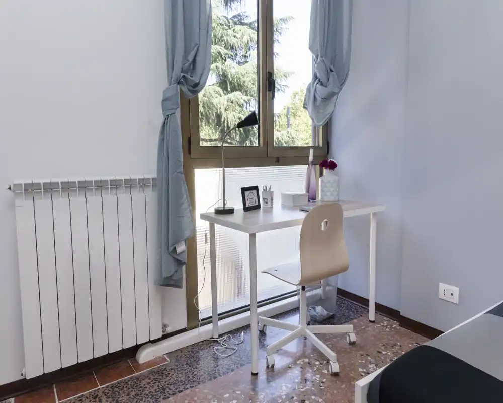 Coin de chambre avec un bureau compact près d'une grande fenêtre laissant entrer une lumière naturelle abondante ; chaise en bois minimaliste, lampe et petits objets décoratifs pour un espace de travail soigné et fonctionnel.