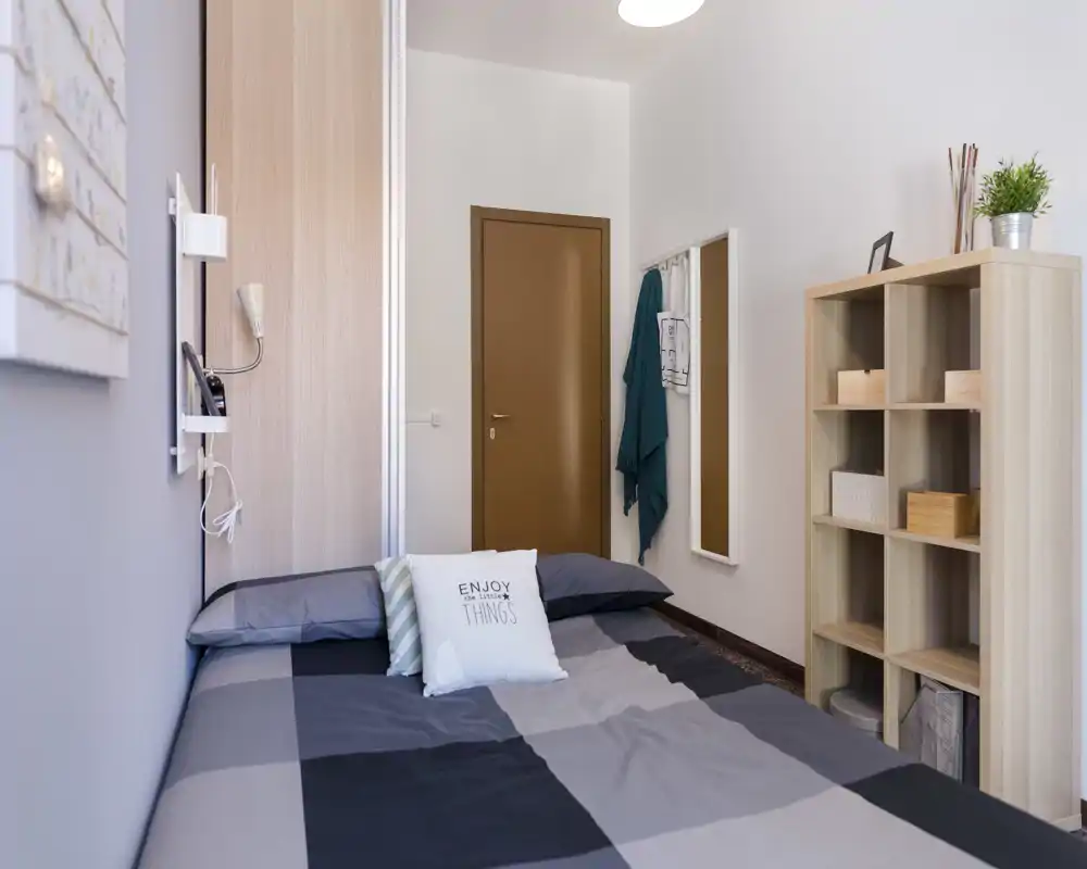 Chambre compacte et soignée avec un lit double en couvre-lit gris à carreaux, coussins décoratifs, une étagère à compartiments et un miroir mural ; lumineuse et bien rangée.