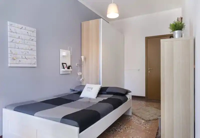 Camera da letto luminosa e ordinata con struttura del letto bianca moderna, parete grigio-blu, armadio e decorazioni essenziali; perfetta per annunci di affitto che valorizzano un design pulito e contemporaneo.