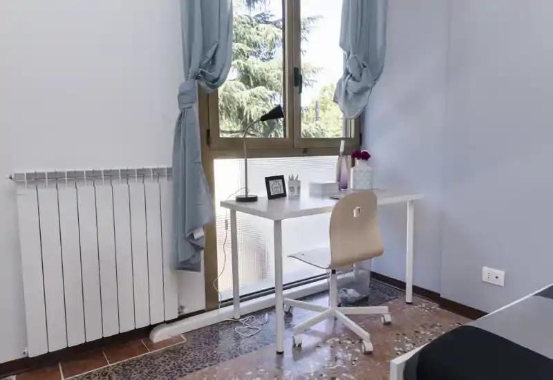 Angolo di camera compatto con una scrivania semplice vicino a una grande finestra, tende chiare e una piccola sedia; visibile parzialmente il letto sul bordo destro.