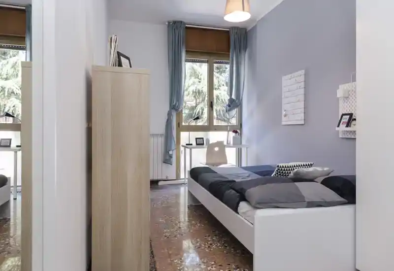 Camera da letto luminosa e ordinata con letto singolo, scrivania vicino alla finestra e accenti blu-grigio che creano un'atmosfera tranquilla.