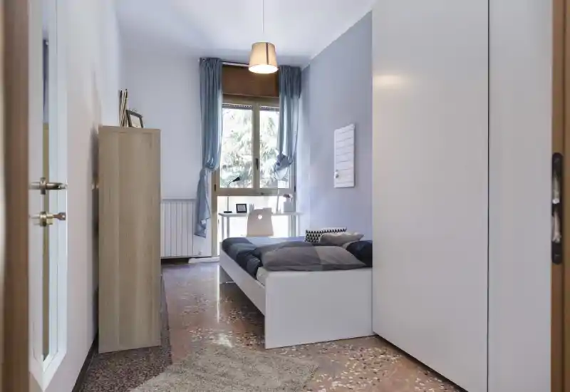 Camera da letto singola luminosa e ordinata con letto bianco, scrivania vicino alla finestra e tende azzurre che lasciano entrare luce naturale.