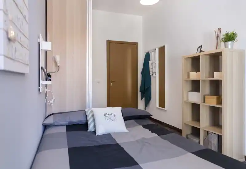 Camera da letto accogliente e ben arredata con letto matrimoniale coperto da una piuma grigia a quadri, cuscini decorativi e uno scaffale in legno per riporre oggetti e piante.