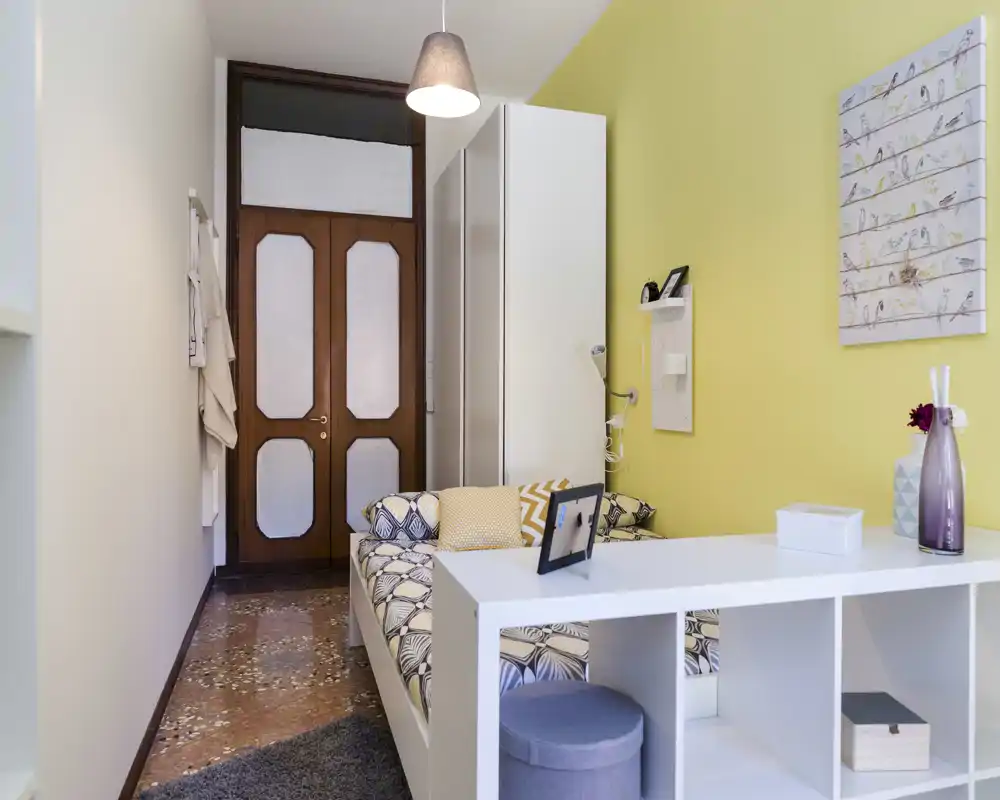 Bright, compact bedroom with a patterned sofa-bed, decorative cushions and a cheerful yellow accent wall; practical shelving separates the sleeping area and adds storage.