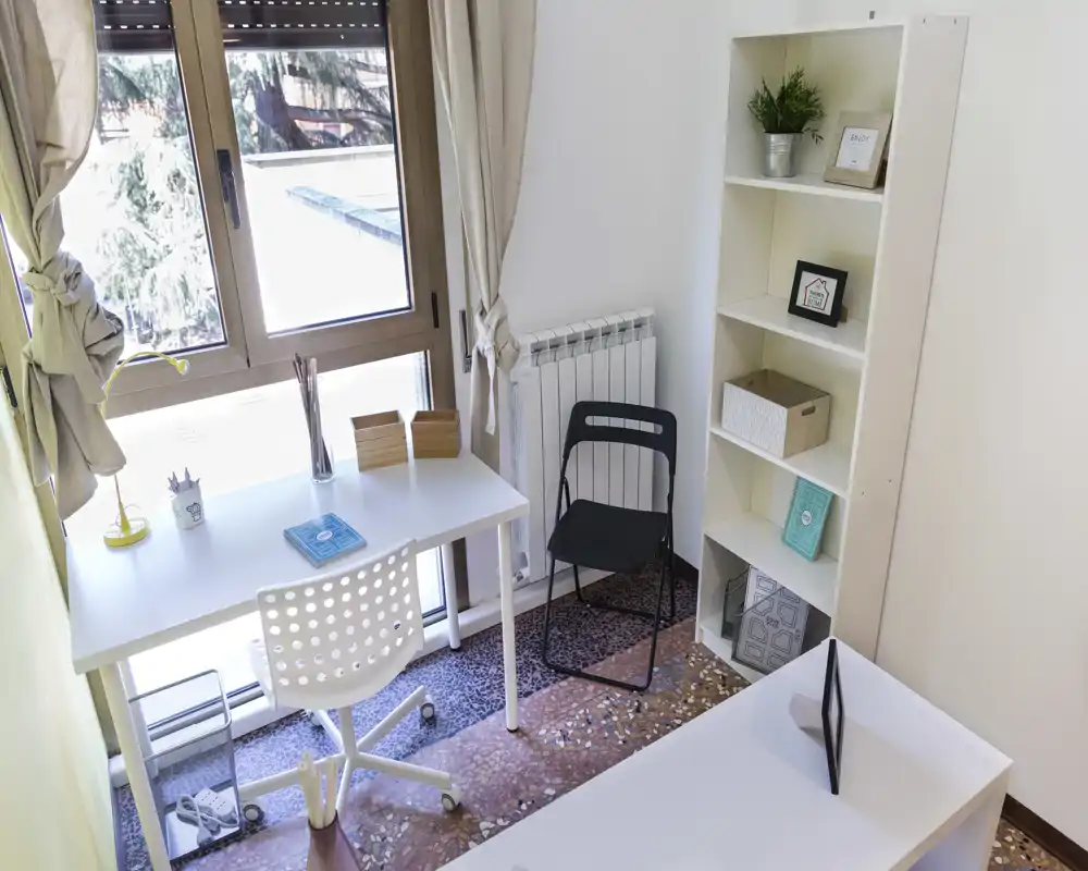 Bright, tidy living area set up as a small home office with a desk by a large window, shelving unit and chairs — ideal for remote work or study.