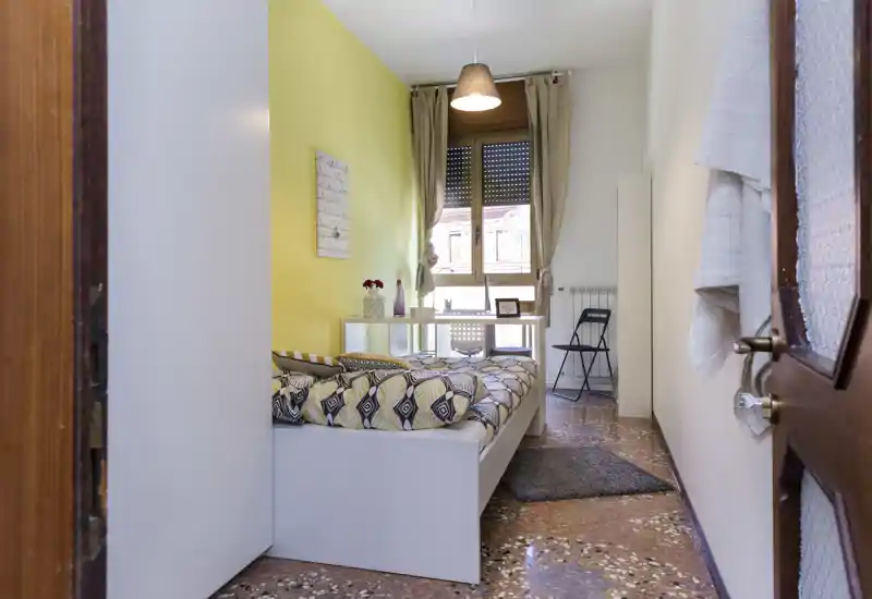 Bright, neatly staged bedroom with a patterned double bed, a yellow accent wall and a window providing natural light; compact desk and chair sit by the window.