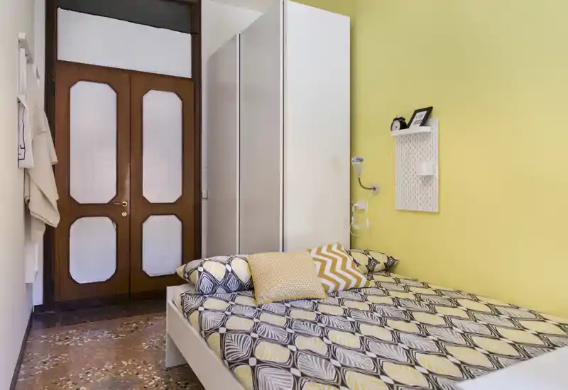 Bright, cozy single bedroom with a patterned bed, decorative cushions and a yellow accent wall. A slim wardrobe and bedside lamp complete the compact, well-styled space.
