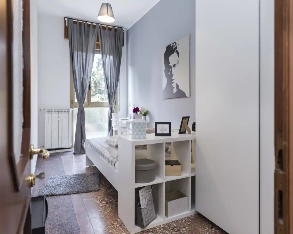 Camera da letto singola luminosa e ben allestita con letto ordinato, contenitore a piede letto e finestra con tende leggere che filtrano la luce naturale.