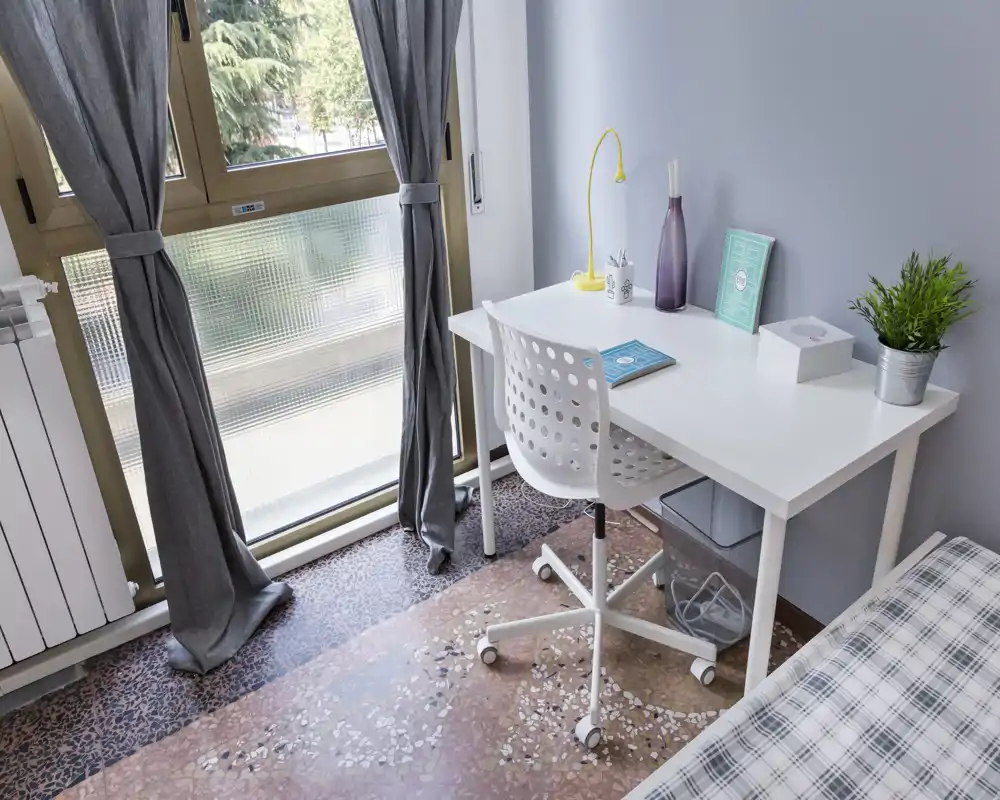 Angolo di camera luminoso e ordinato con scrivania bianca e sedia accanto al letto a quadri. La luce naturale entra dalle grandi finestre con tende grigie, creando un'area moderna per studio e riposo.