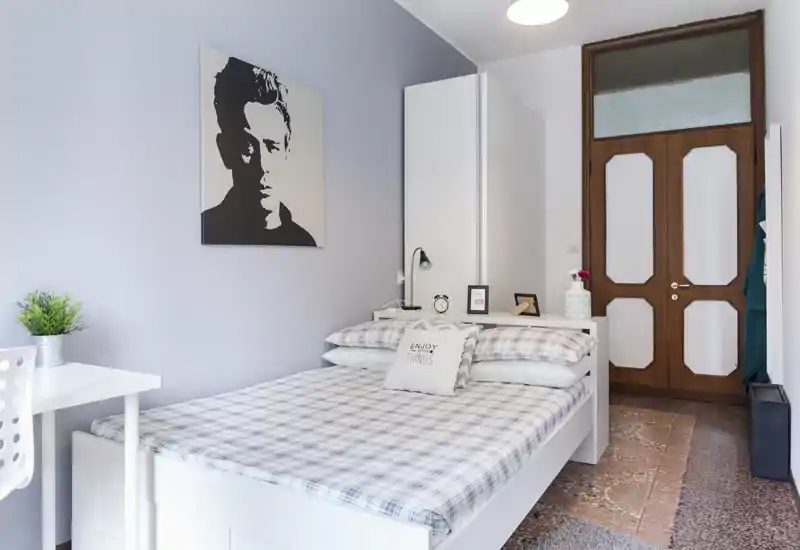 Dormitorio luminoso y ordenado con cama completa vestida con ropa de cama estampada, mesita con lámpara y pequeños objetos decorativos, y un armario alto junto a la puerta. La habitación transmite sensación acogedora con luz suave y clara.