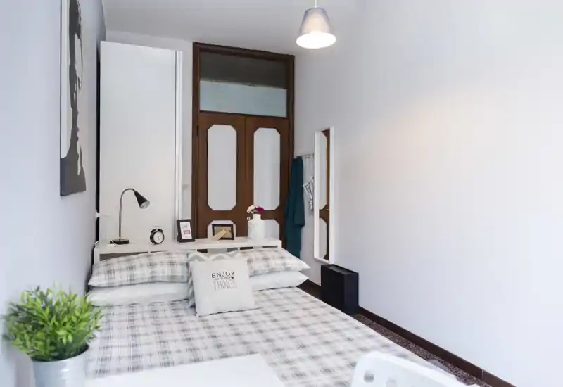 Dormitorio luminoso y ordenado con cama doble de ropa a cuadros, mesita con lámpara y decoración, y paredes blancas que crean un ambiente moderno y luminoso.