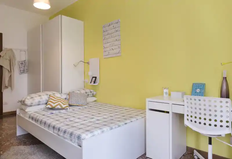 Dormitorio luminoso y ordenado con cama, escritorio y armario frente a una pared amarilla alegre; perfecto para una estancia confortable.