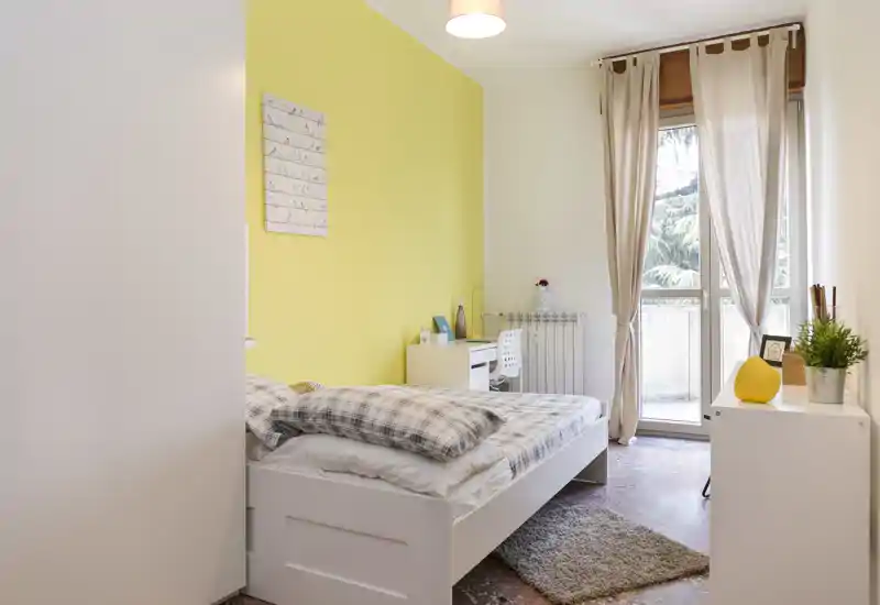 Dormitorio individual luminoso y acogedor con cama blanca, pared amarilla y puerta balcón que aporta luz natural. Pequeño escritorio y almacenamiento, adecuado para un estudiante o una persona sola.