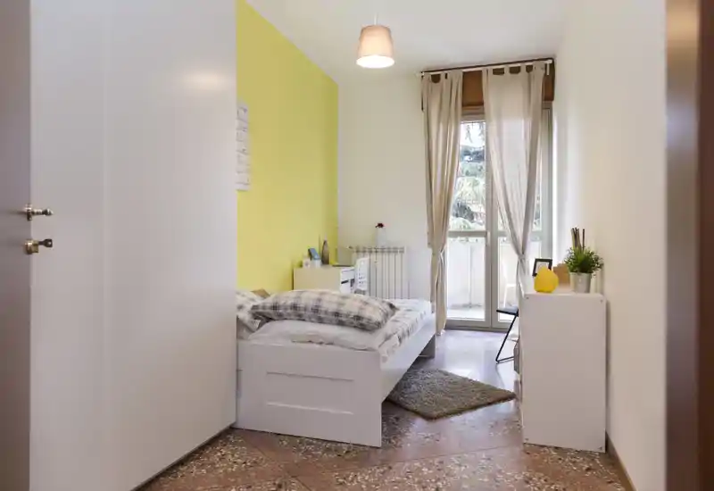 Dormitorio luminoso y ordenado con una cama individual blanca, ropa de cama a cuadros y una puerta a balcón que aporta luz natural. La pared amarilla y la decoración sencilla crean un ambiente fresco y acogedor.