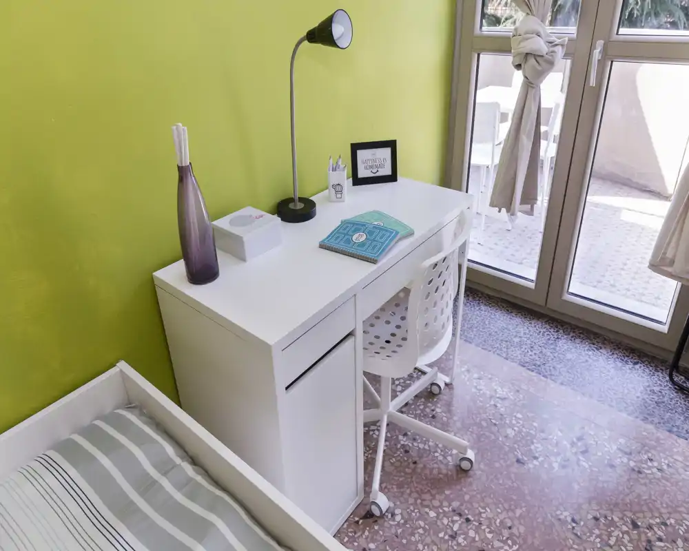 Bright, tidy bedroom corner with a single bed and a neat white desk positioned by a glass door leading to a small balcony. The space features modern minimal decor, natural light and a functional workspace ideal for studying or remote work.