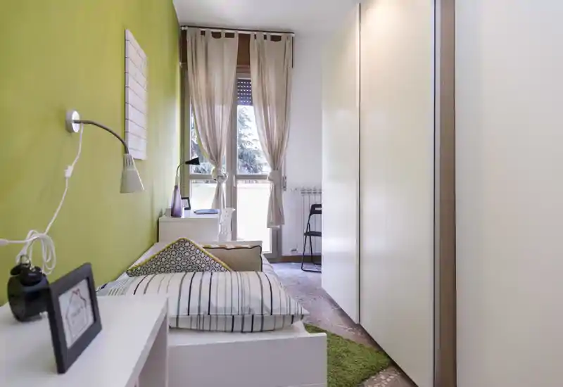 Dormitorio individual ordenado con pared verde, cama hecha, luz natural desde el balcón y armario práctico a la derecha.