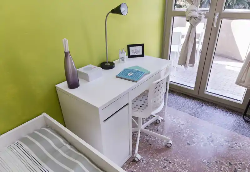 Rincón de dormitorio luminoso y ordenado con cama individual y un escritorio blanco junto a una puerta de cristal que da a un pequeño balcón. Decoración moderna y minimalista, luz natural y zona de trabajo funcional.