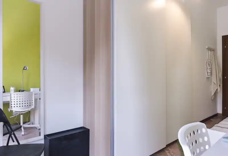 Vista de dormitorio compacto con armario blanco, borde de la cama parcialmente visible y un pequeño escritorio reflejado en el espejo; luminoso y mobiliario sencillo.