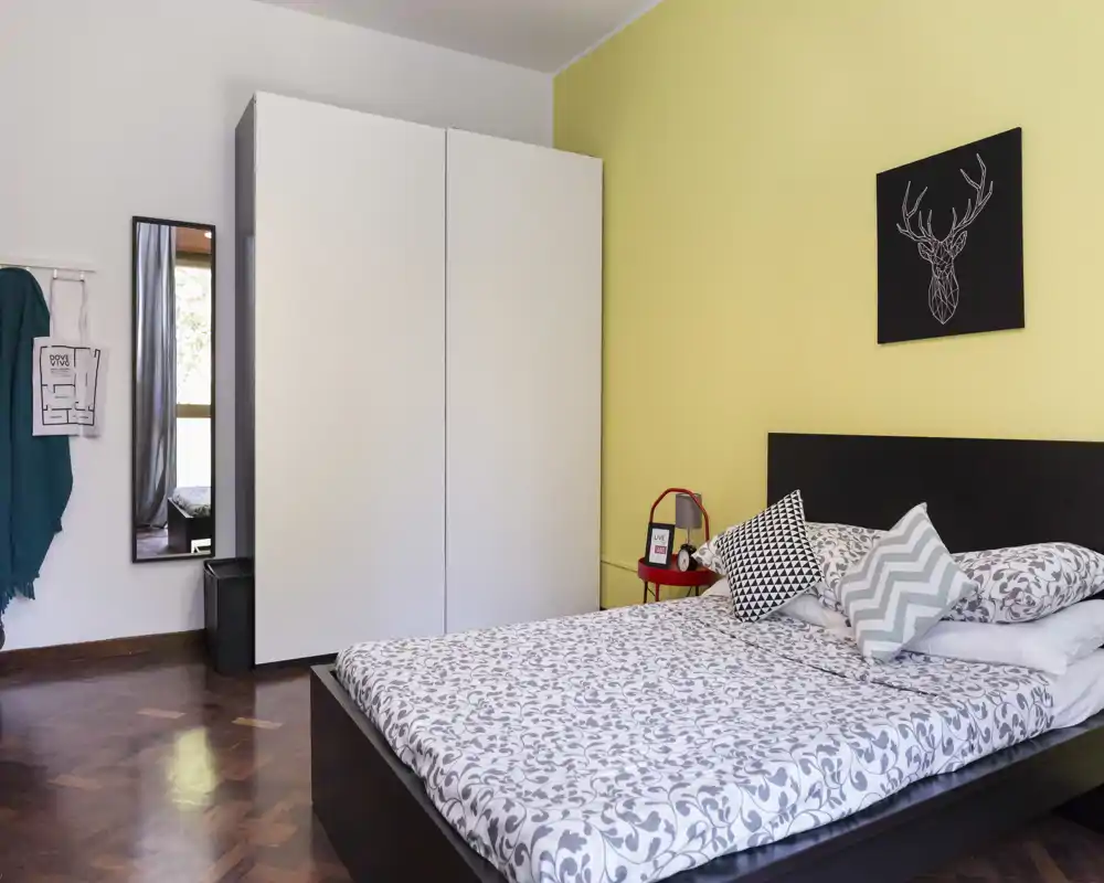 Camera da letto ben arredata con letto matrimoniale, biancheria a motivo, cuscini decorativi e un armadio minimale davanti a una parete d'accento gialla.