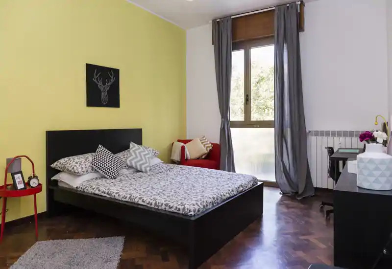 Camera da letto luminosa e ben arredata con letto matrimoniale, biancheria a fantasia, cuscini decorativi e una piccola poltrona rossa vicino alla finestra; parquet in legno e tessili creano un'atmosfera accogliente.