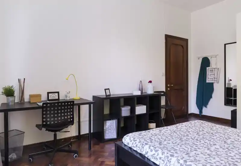 Camera da letto luminosa e ordinata con letto matrimoniale dal copriletto a fantasia, scrivania con sedia e scaffale aperto — ideale per riposo e lavoro da remoto.