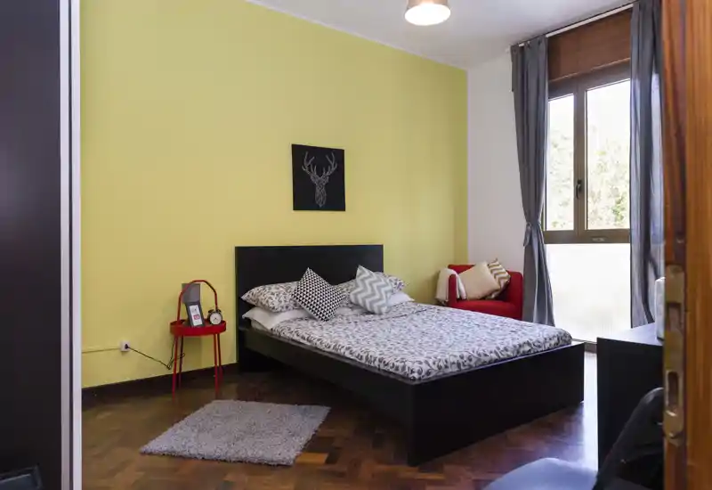 Camera da letto luminosa e ben arredata con letto matrimoniale contro una parete gialla, cuscini decorativi, un piccolo comodino e una poltrona accogliente vicino alla finestra.