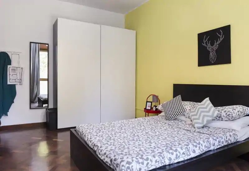 Camera da letto ben arredata con letto matrimoniale, biancheria a motivo, cuscini decorativi e un armadio minimale davanti a una parete d'accento gialla.