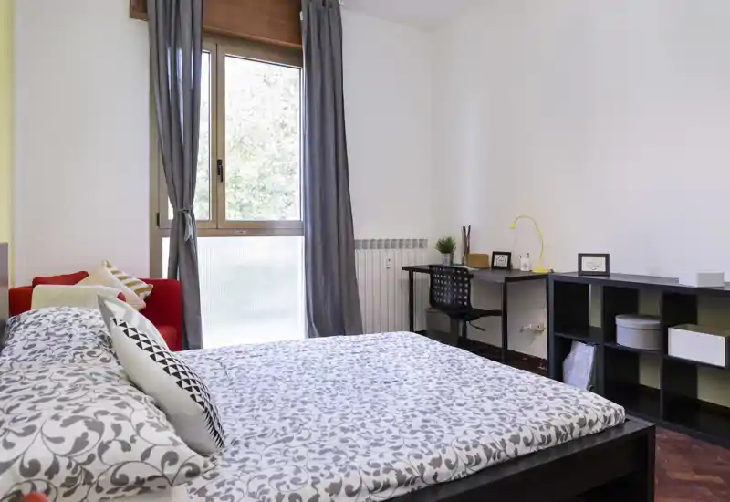 Camera da letto luminosa e ben allestita con letto matrimoniale a fantasia, scrivania e scaffalatura — ideale per un soggiorno confortevole con luce naturale da una grande finestra.